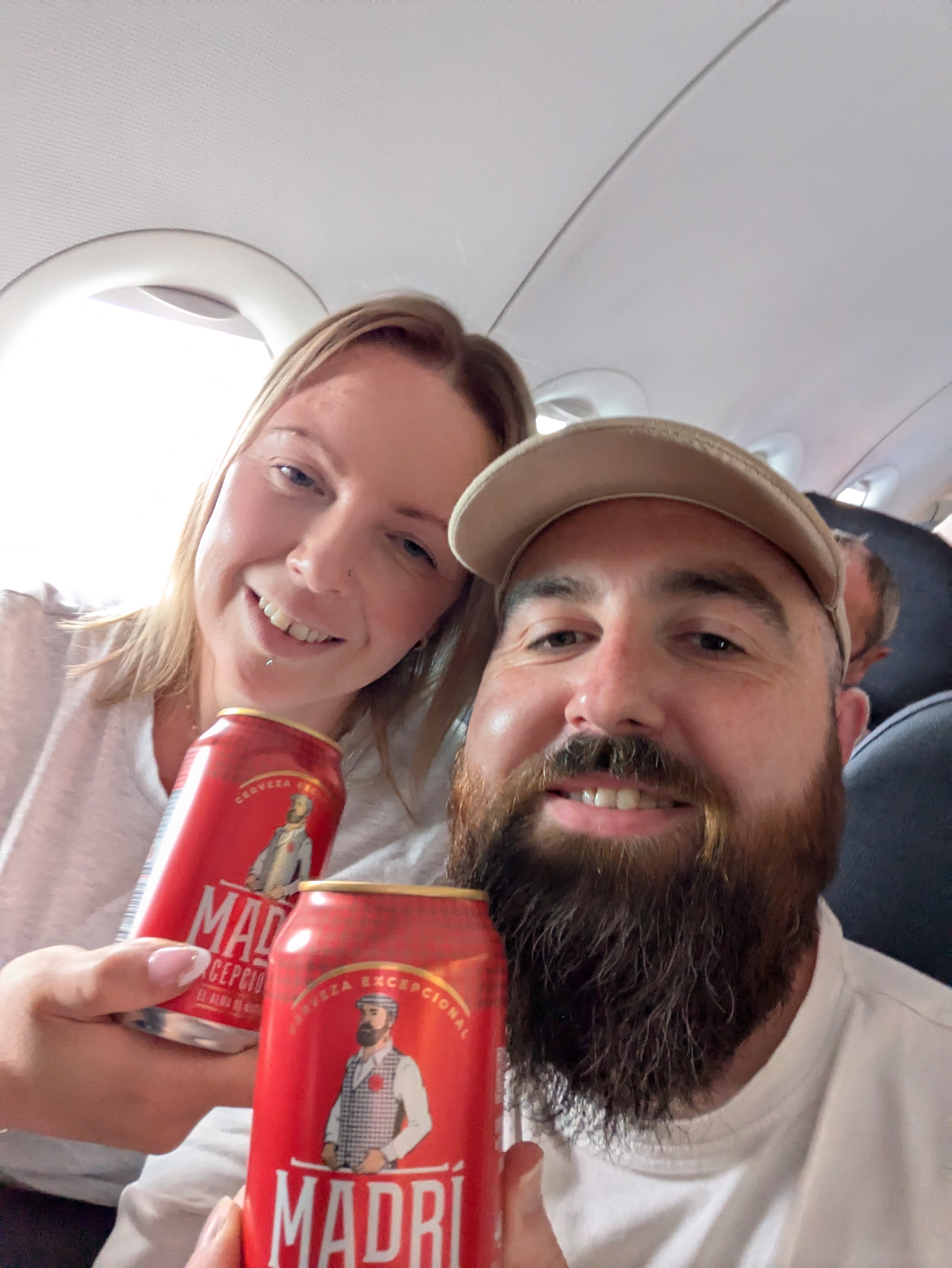 A couple on an aeroplane, smiling and holding cans of Madri beer, with the aircraft interior visible in the background.